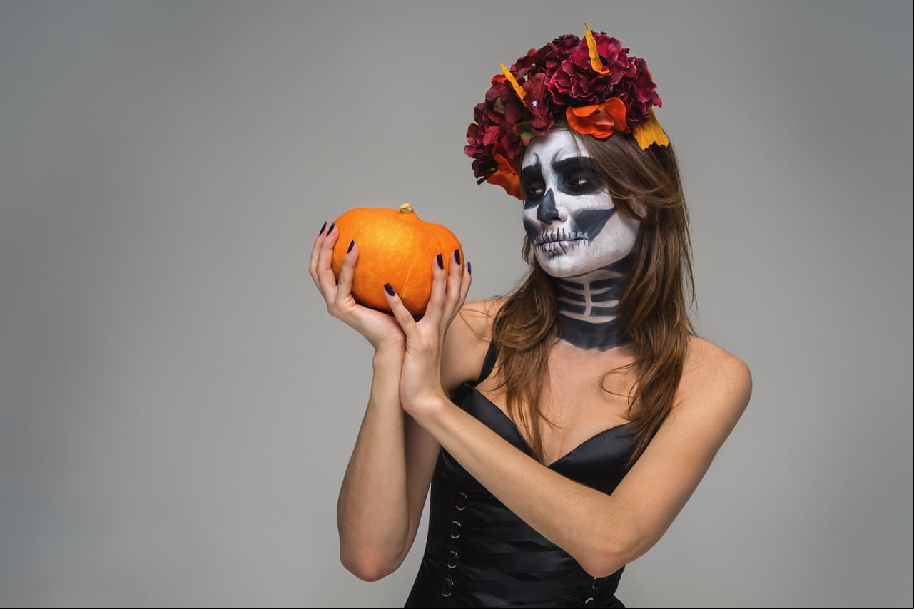 Portrait of young beautiful girl with fearful halloween skeleton makeup with a wreath Katrina Calavera made of flowers on her head, holding pumpkin over gray background