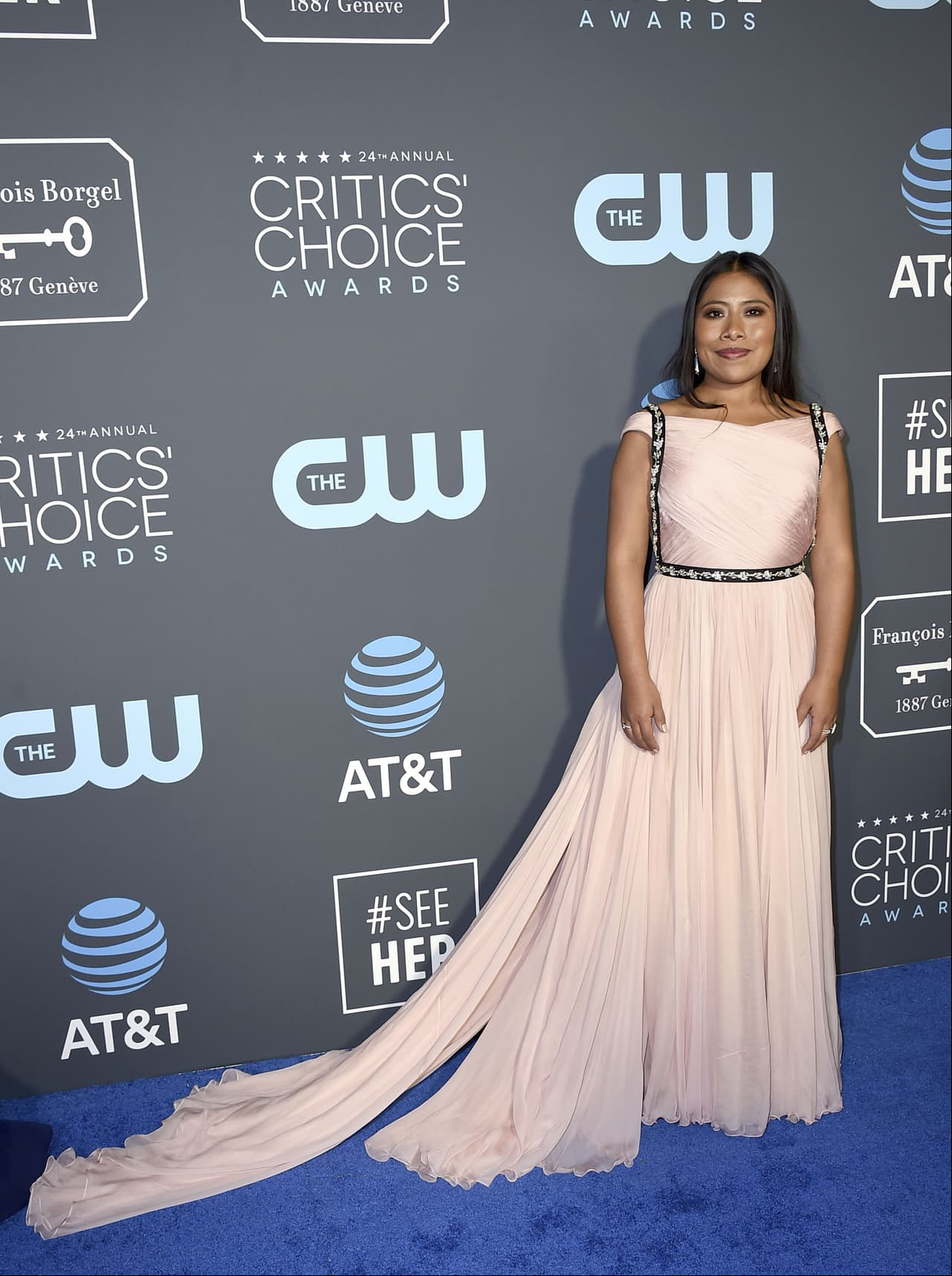 En la alfombra roja de los Critics’ Choice Awards, Yalitza lució un vestido Prada de color rosa claro con manga a la altura de los hombros, resaltando los detalles negros con brillantes; además usó unos pendientes plateados y tres anillos del mismo tono.